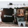 Black ripstop transport bag with reinforced construction and adjustable shoulder strap next to a compact metal outdoor stove, suitable for camping, farm, and garden use.