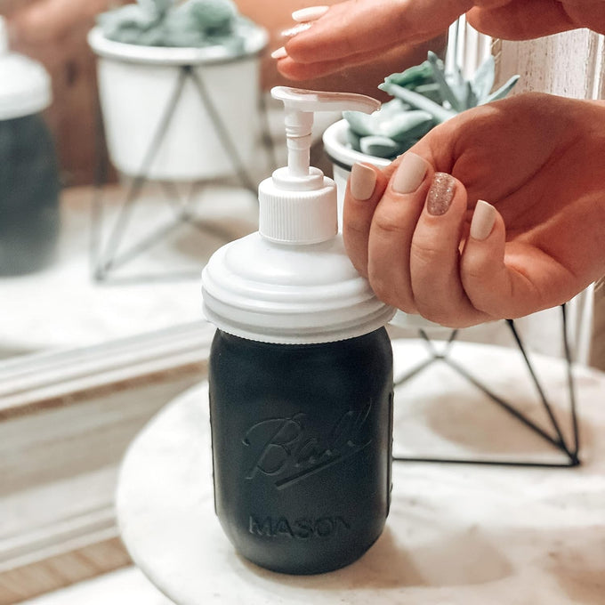 Hand pressing white pump lid on black mason jar filled with liquid, set on marble countertop with decorative plants in background, part of USA Made, Home & Decor, Household Helpers, Best Sellers, Promo Eligible collections.