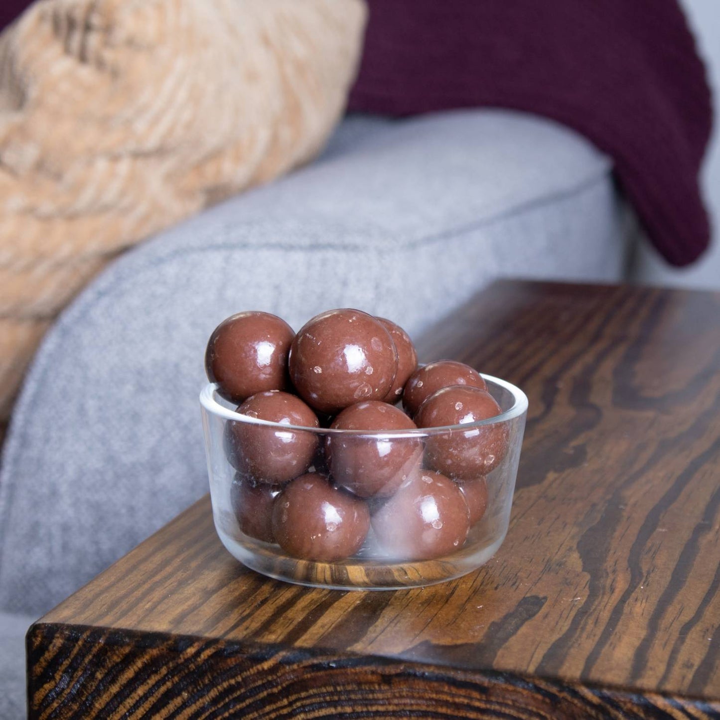 Clear glass bowl filled with glossy milk chocolate malted milk balls set on a dark wood table in a cozy living room, representing snacks from Kitchen & Food, Candy, and USA Made collections.