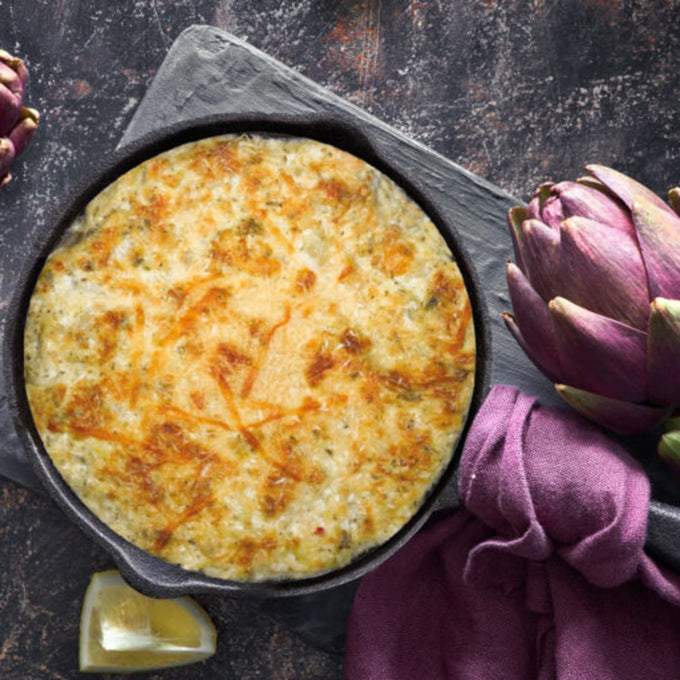 Golden baked dip with melted cheese and herbs in an 8-inch cast iron skillet, styled with fresh artichoke, lemon wedge, and purple cloth, suited for Kitchen & Food, Dry Goods and Mixes, Best Sellers collections.