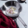 Handcrafted fully clad stainless steel fry pan with a riveted handle containing sliced shallots and garlic, styled with a red cloth, featured in Kitchen & Food, Cookware, Pots and Pans, Best Sellers, and Promo Eligible collections.