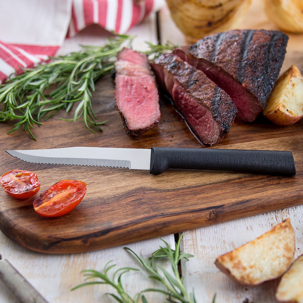 Handcrafted serrated steak knife with high carbon surgical stainless steel blade and black handle on wooden board with sliced grilled steak, roasted potato, tomato, and fresh rosemary, ideal for Kitchen & Food, Farm, Garden & Tool, and USA Made collections.