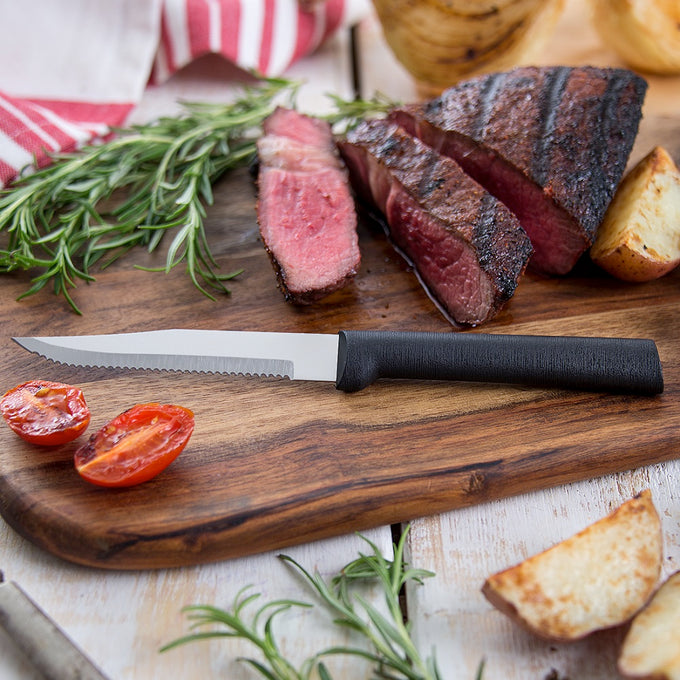 Handcrafted serrated steak knife with high carbon surgical stainless steel blade and black handle on wooden board with sliced grilled steak, roasted potato, tomato, and fresh rosemary, ideal for Kitchen & Food, Farm, Garden & Tool, and USA Made collections.