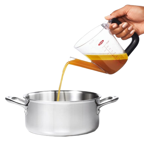 Clear plastic fat separator with a black cushioned grip handle and red stopper, pouring liquid from the spout into a stainless steel pot, ideal for Kitchen & Food and Cooking Helpers collections.