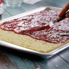 Close-up of a textured baking pan with a light cake base being spread with red jam using a wooden-handled spatula, perfect for Kitchen & Food, Cookware, Baking Supplies, and Pots and Pans collections.
