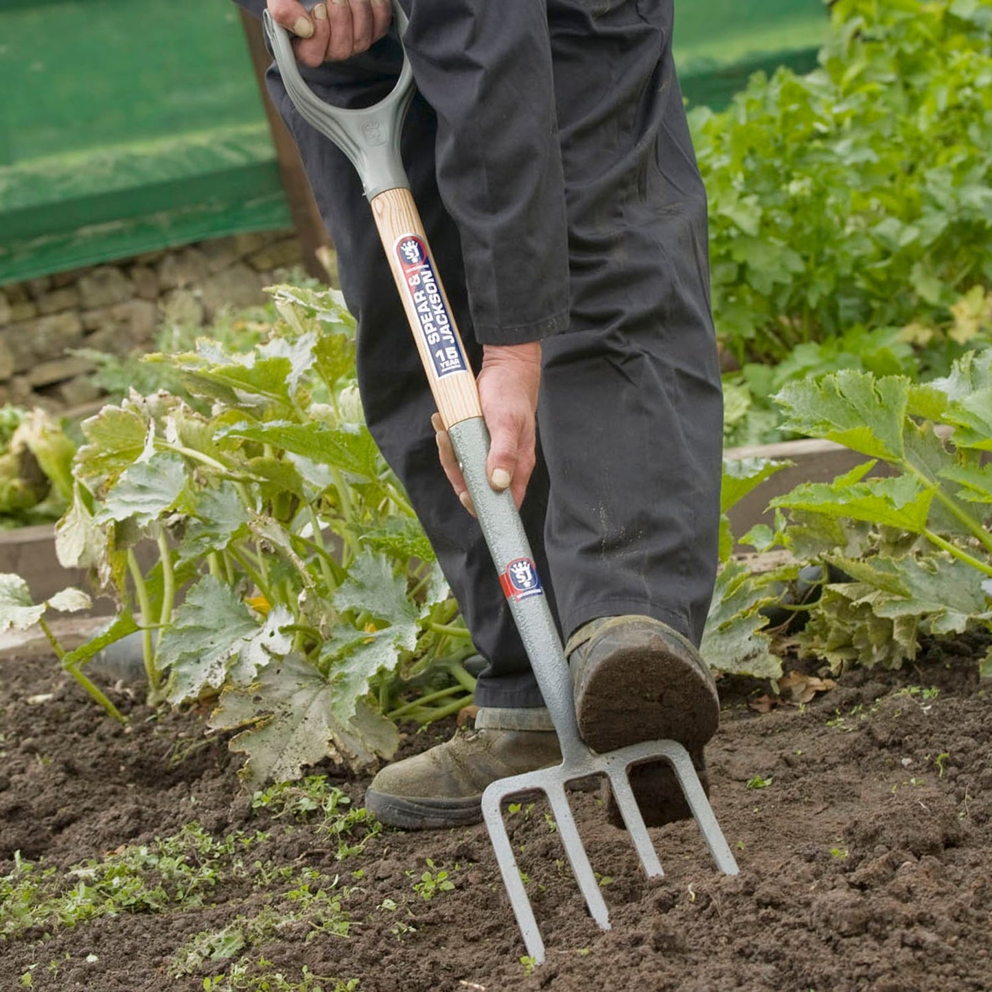 Person using a Spear & Jackson Neverbend Carbon Digging Fork with forged carbon steel tines and Supergrip handle to loosen soil in a garden bed, suitable for farm, garden, and lawncare tasks.