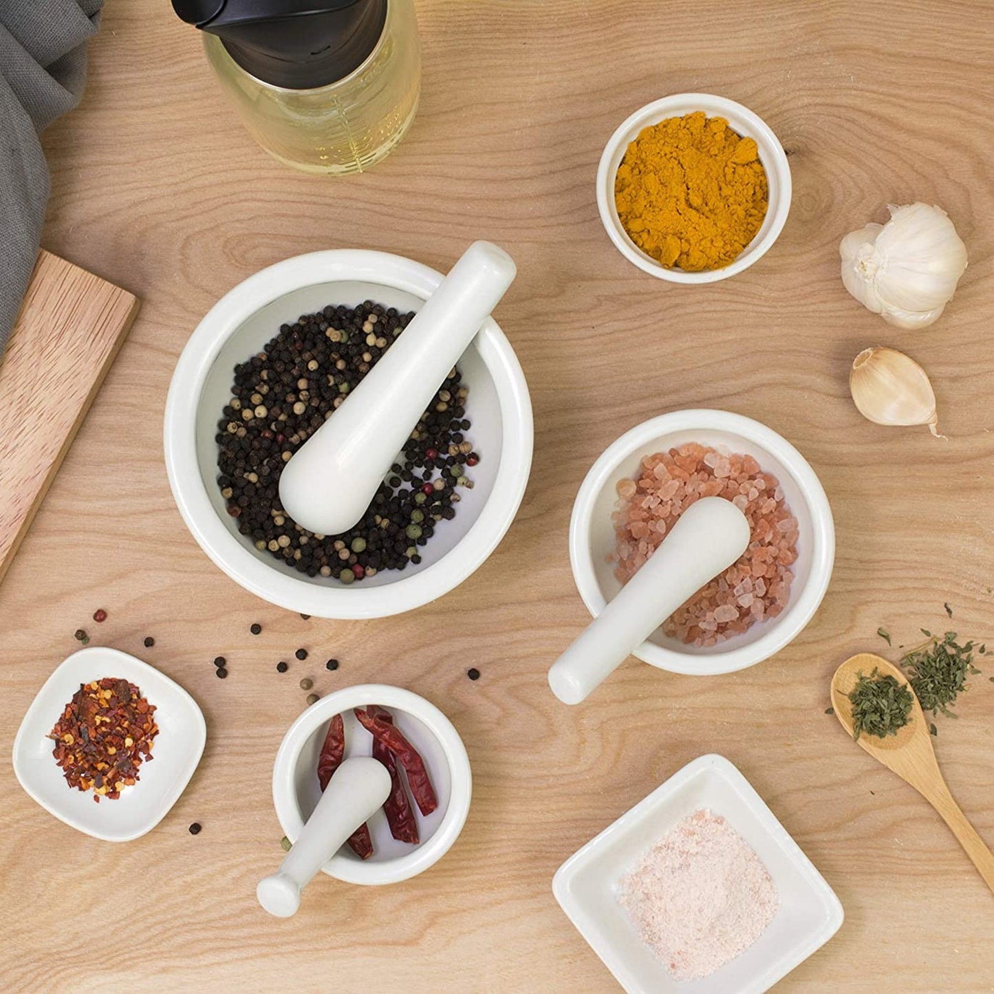 Set of three white porcelain mortars and pestles in different sizes, filled with whole peppercorns, pink salt, and dried chilies, displayed on wooden surface with spices, ideal for kitchen food prep and cooking helpers.