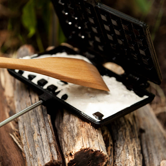 Cast iron waffle iron with a wooden handle filled with waffle batter, set on a rustic log outdoors, ideal for Farm, Garden & Tool, Camping, Best Sellers, and Promo Eligible collections.