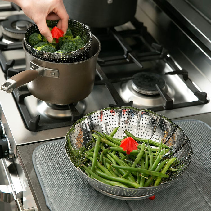 Hand placing broccoli inside a collapsible stainless steel vegetable steamer basket with red umbrella-shaped handle set over a pot on gas stove, with green beans in similar basket on stovetop cover, ideal for Kitchen & Food, Cookware, Pots and Pans, Kitchen Gadgets, Cooking and Baking Helpers, Best Sellers, Promo Eligible collections.