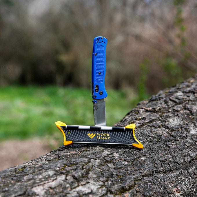 Outdoor scene with a blue-handled folding knife held upright by a compact Work Sharp Pocket Knife Sharpener with black body and yellow corner grips, featured for Farm, Garden & Tool and Knives & Axes collections.