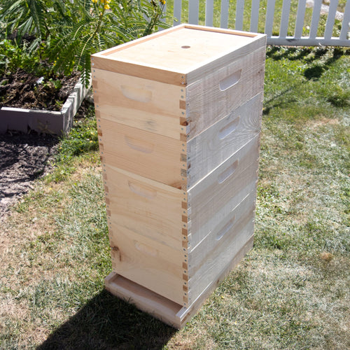 Stack of unfinished wooden beehive boxes standing outdoors on grass near garden bed and white fence, handcrafted in the USA, Amish made, ideal for farm, garden, and beekeeping supplies collections.