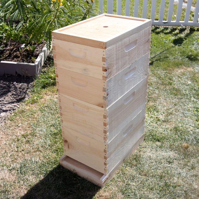 Stack of unfinished wooden beehive boxes standing outdoors on grass near garden bed and white fence, handcrafted in the USA, Amish made, ideal for farm, garden, and beekeeping supplies collections.