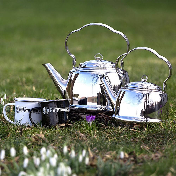 Two polished stainless steel teakettles with elegant curved handles sitting on grass with blooming flowers, accompanied by enamel mugs, perfect for Farm, Garden & Tool, Kitchen & Food, Camping, Brewing Coffee and Tea, Best Sellers, and Promo Eligible collections.
