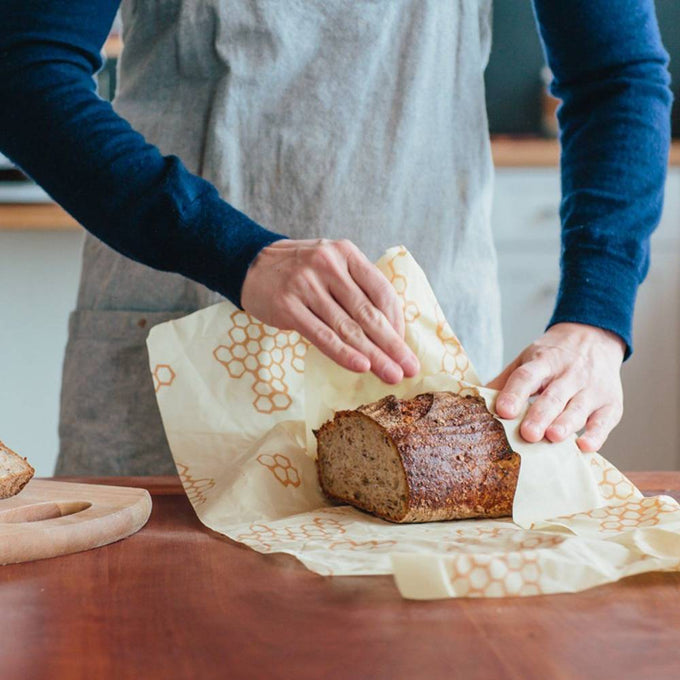 Hands wrapping a loaf of bread in a reusable beeswax muslin cloth wrap with honeycomb pattern, handcrafted in the USA, ideal for Kitchen & Food and Bread Making Supplies collections.
