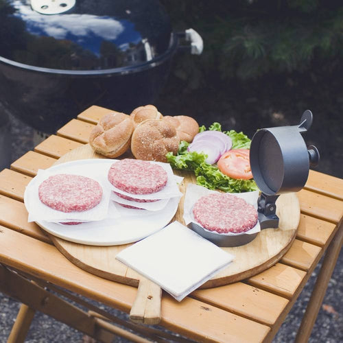 Black metal single hamburger press with adjustable thickness lever, shown with raw beef patties on wax paper, fresh burger buns, lettuce, tomato, and onion on wooden table, ideal for Kitchen & Food, Specialty Equipment, Butchering Supplies, Best Sellers, and Promo Eligible collections.