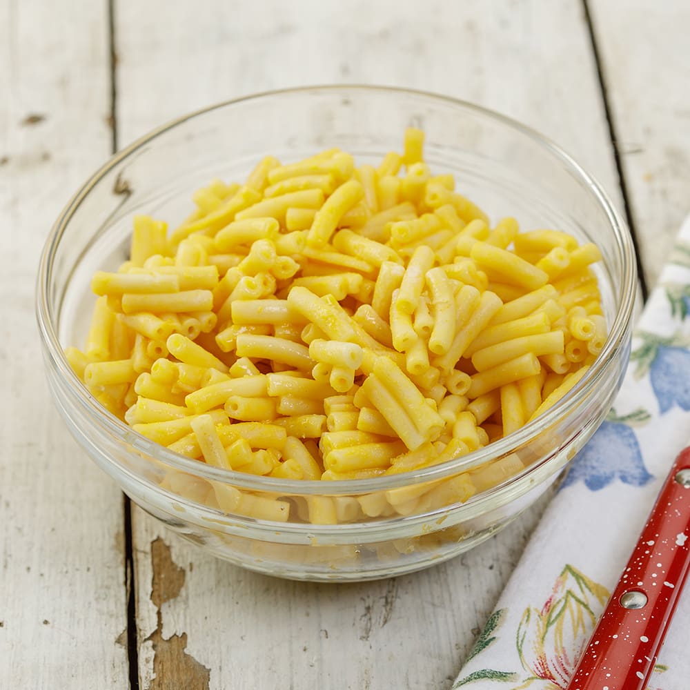 Clear glass bowl filled with creamy prepared macaroni and cheese on a rustic wooden surface next to a floral napkin and red-handled utensil, perfect for pantry and Kitchen & Food collections.