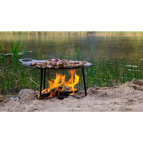 Steel griddle and fire bowl with three-legged design cooking meat over an open flame by a lakeside, ideal for outdoor use in Farm, Garden & Tool, Camping, Best Sellers, and Promo Eligible collections.