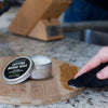 Small metal tin of cutting board wax conditioner with black label opened on a kitchen countertop while a hand applies wax to a round wooden cutting board, featured in Kitchen & Food and Food Prep collections.