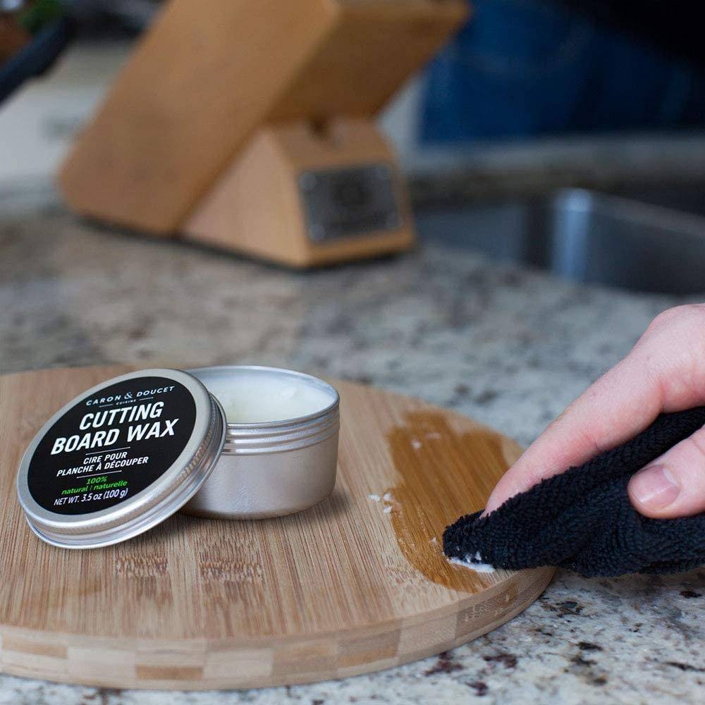 Small metal tin of cutting board wax conditioner with black label opened on a kitchen countertop while a hand applies wax to a round wooden cutting board, featured in Kitchen & Food and Food Prep collections.