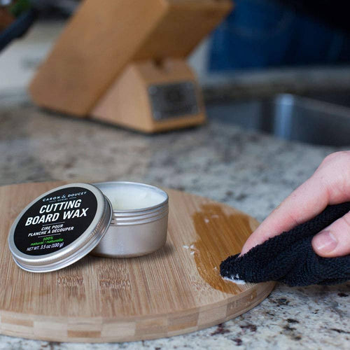 Small metal tin of cutting board wax conditioner with black label opened on a kitchen countertop while a hand applies wax to a round wooden cutting board, featured in Kitchen & Food and Food Prep collections.