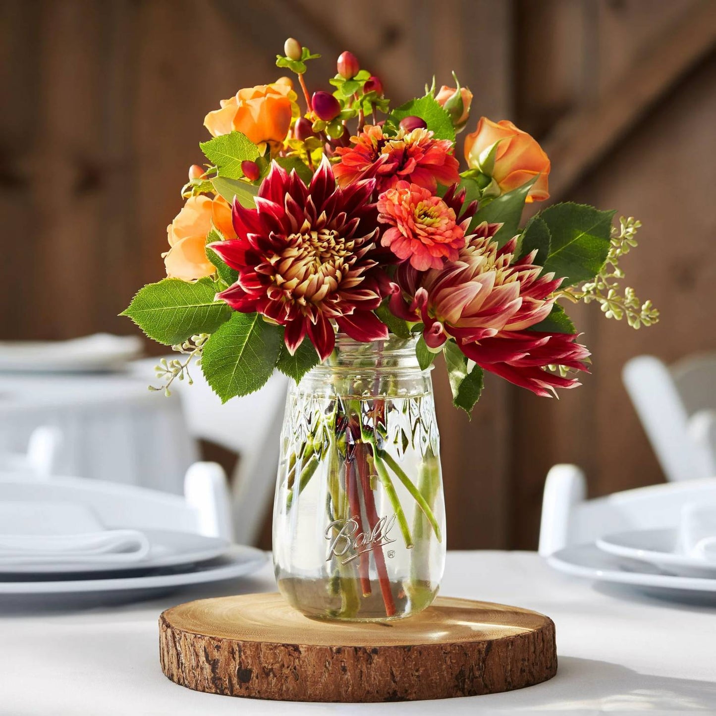 Clear regular-mouth pint jar with an engraved diamond pattern filled with autumn-colored flowers, set on a rustic wood slice, ideal for Kitchen & Food and Storage collections.