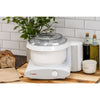 Compact white stand mixer with clear lid on wooden countertop surrounded by bread, oil bottles, salt container, and greenery, perfect for baking and kitchen helpers collections.