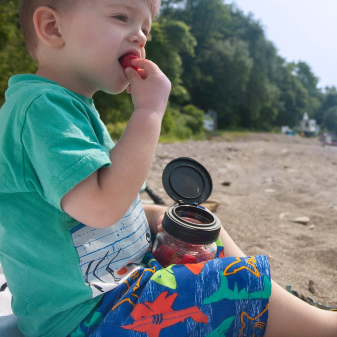 Young child sitting outdoors near a lakeshore, eating a snack from a glass jar with a black flip top lid, showcasing reusable kitchen accessories in Home & Decor and Kitchen Gadgets collections.