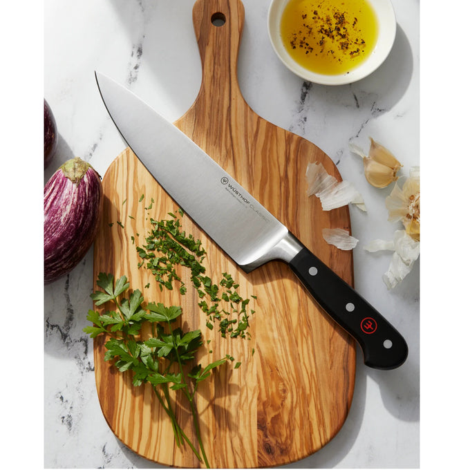 Sharp chef's knife with high carbon stainless steel blade and triple-riveted full tang handle resting on a wooden cutting board with chopped herbs, garlic, and olive oil, ideal for Kitchen Knives, Food Prep, and Best Sellers collections.