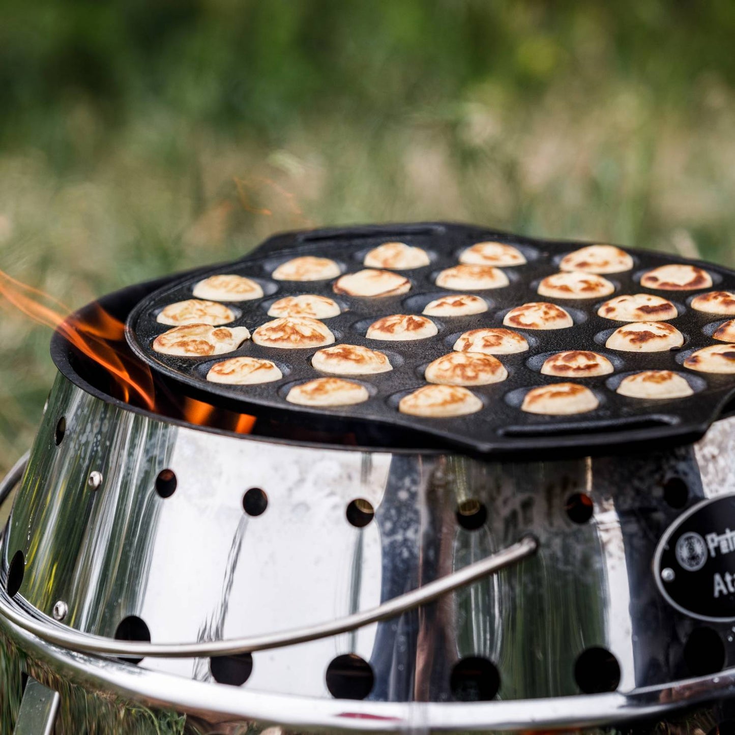Cast iron pan with 30 mini Dutch pancakes cooking over an open fire on a stainless steel camp stove, designed for outdoor use in camping, kitchen, and cookware collections.
