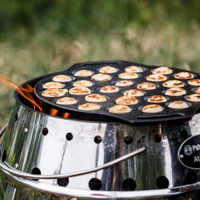 Cast iron pan with 30 mini Dutch pancakes cooking over an open fire on a stainless steel camp stove, designed for outdoor use in camping, kitchen, and cookware collections.