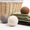 Set of three all-natural wool dryer balls in brown, gray, and off-white colors arranged near folded green towels and woven laundry basket, crafted for Laundry Supplies and Home & Decor collections.