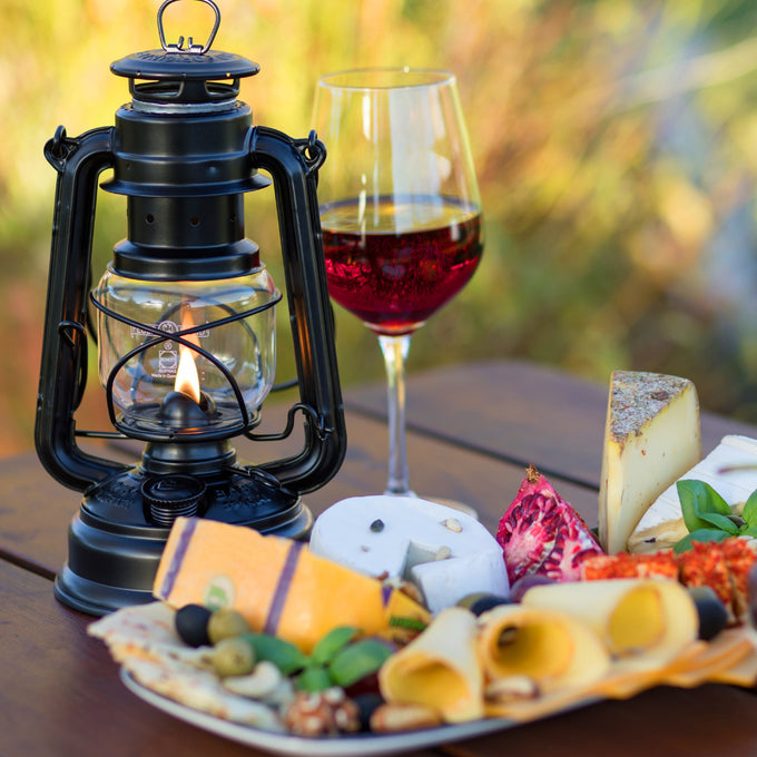 Handcrafted black galvanized oil lantern with thermal heat-resistant SUPRAX globe, glowing warmly on a wooden table next to a charcuterie plate and glass of red wine, ideal for camping, emergency supplies, and outdoor use in Lamps & Lighting collections.