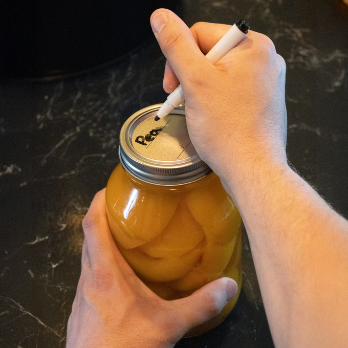 Person writing a label on a silver-toned regular-mouth canning lid with BPA-free sealing compound placed on a jar of preserved peaches, suitable for Kitchen & Food and Food Preservation collections.