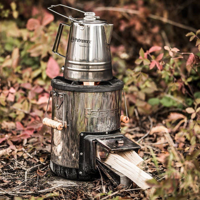 Petromax Rocket Compact outdoor stove made of steel with wooden side handles, burning wood logs to heat a stainless steel kettle, set outdoors in a natural camping area, featured in Farm, Garden & Tool and Camping collections.