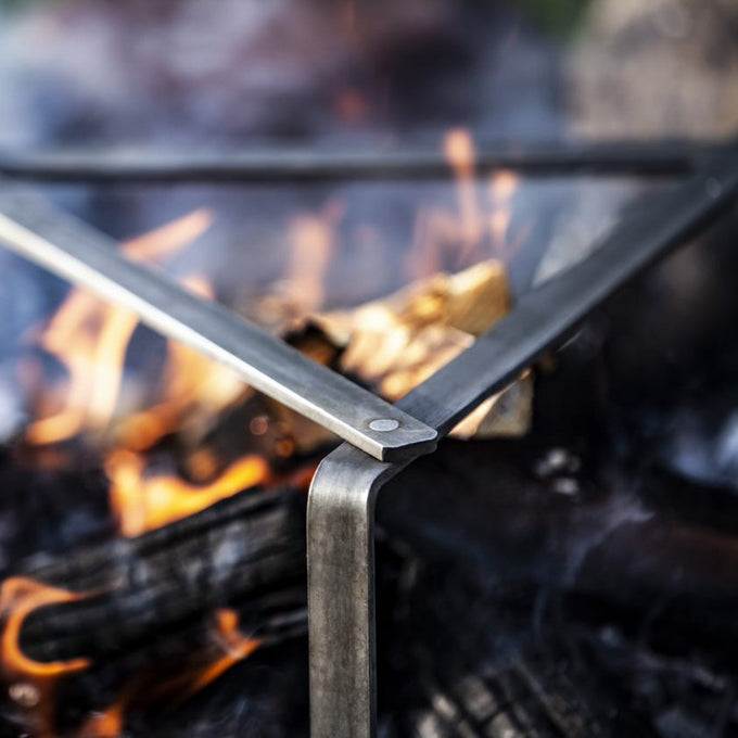 Close-up of a sturdy steel cooking stand leg positioned over glowing campfire logs, ideal for supporting heavy cookware during outdoor cooking, featured in Farm, Garden & Tool and Camping collections.