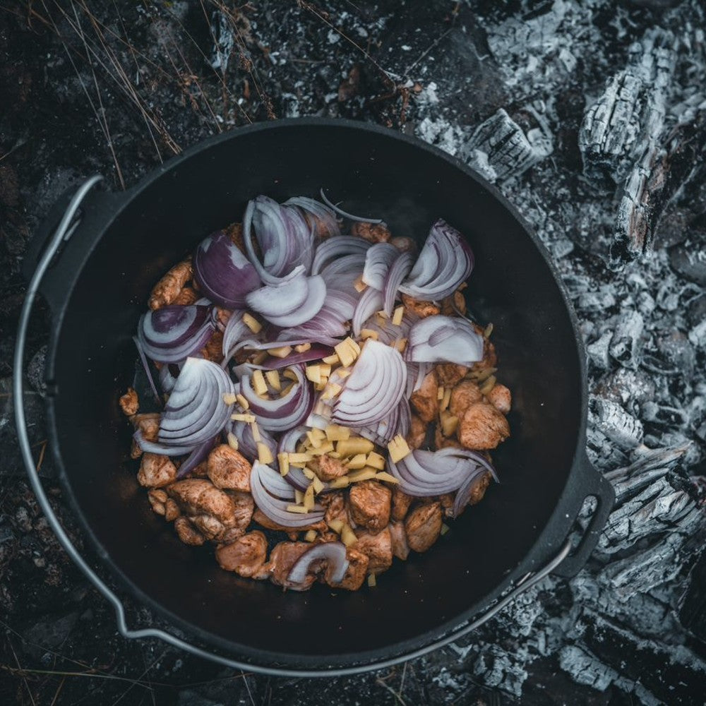 Open cast iron Dutch oven with flat base containing seasoned chicken pieces, sliced red onions, and chopped garlic cooking over glowing embers, ideal for Kitchen & Food, Cookware, Cast Iron, Best Sellers, Promo Eligible collections.