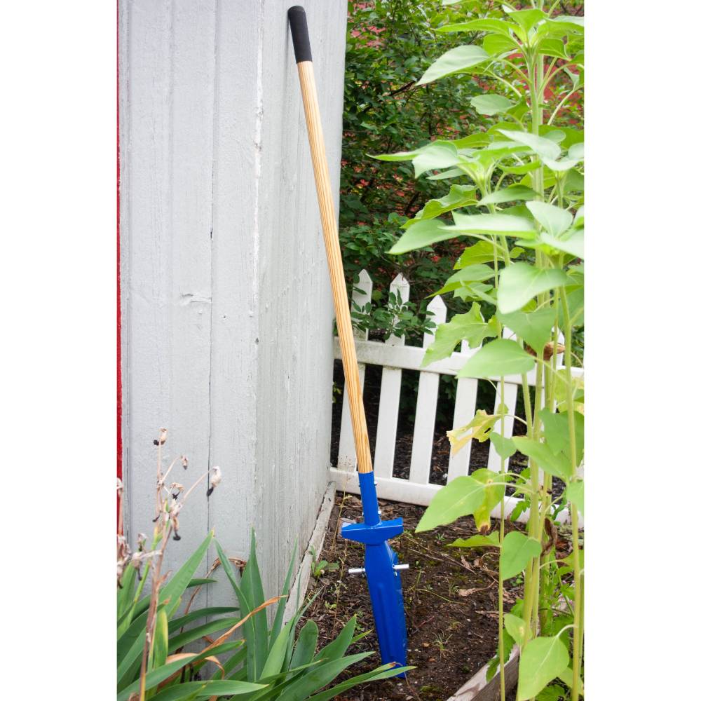Narrow blue trenching shovel with a long wooden handle and black grip, positioned in soil beside a white fence and garden plants, ideal for farm tools and hand digging tasks.