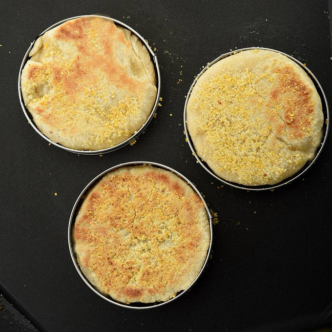 Three English muffins cooking inside stainless steel rings on a griddle, showcasing even browning and cornmeal topping, suitable for Kitchen & Food and Best Sellers collections.