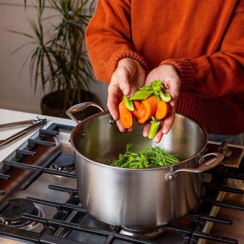 Person adding fresh sliced carrots, snap peas, and herbs into a handcrafted fully clad steel stock pot on a gas stove, featured in Kitchen & Food cookware collections.