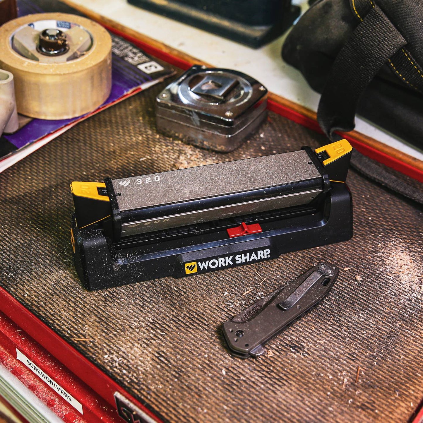 Angle-guided benchstone knife sharpener with coarse and fine abrasive surfaces in black and yellow, shown on a textured mat with a folding knife and workshop tools, ideal for sharpening in Farm, Garden & Tool and Kitchen & Food collections.