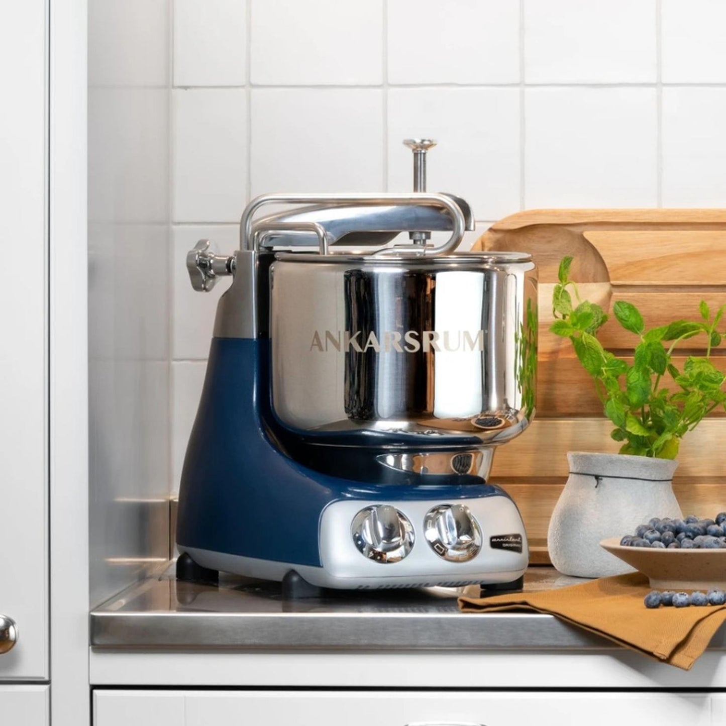 Ankarsrum Assistent stand mixer with polished stainless steel bowl and blue base, placed on a kitchen countertop surrounded by wooden boards and fresh herbs, featured in Kitchen & Food, Cookware, Baking Supplies, Kitchen Gadgets, and Bread Making Supplies collections.