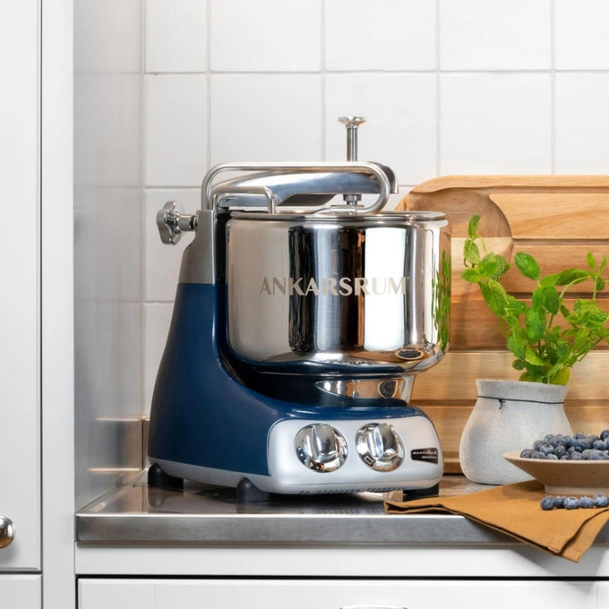 Ankarsrum Assistent stand mixer with polished stainless steel bowl and blue base, placed on a kitchen countertop surrounded by wooden boards and fresh herbs, featured in Kitchen & Food, Cookware, Baking Supplies, Kitchen Gadgets, and Bread Making Supplies collections.
