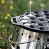 Cast iron poffertjes pan with multiple shallow molds, being filled with batter over an outdoor fire setup, ideal for camping, kitchen, and cast iron cookware collections.