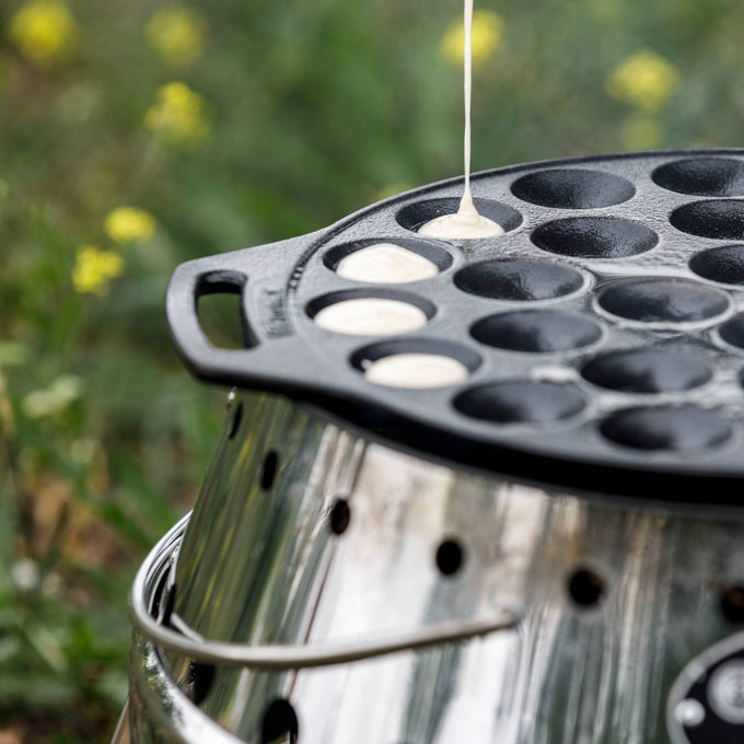 Cast iron poffertjes pan with multiple shallow molds, being filled with batter over an outdoor fire setup, ideal for camping, kitchen, and cast iron cookware collections.