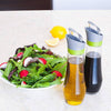 Pair of glass automatic oil bottles with flip-top lids and drip catchers, filled with oil and vinegar, displayed with fresh salad and lemon on countertop in Kitchen & Food and Cookware collections.