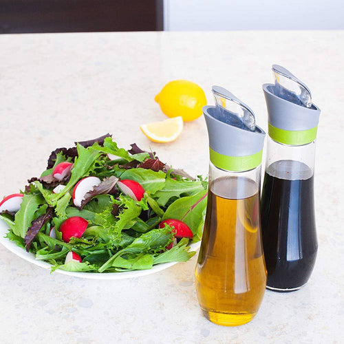 Pair of glass automatic oil bottles with flip-top lids and drip catchers, filled with oil and vinegar, displayed with fresh salad and lemon on countertop in Kitchen & Food and Cookware collections.
