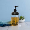Clear glass mason jar filled with golden soap and topped with a black pump lid, sitting on a white surface with folded blue cloths and a small succulent nearby, highlighting eco-friendly, USA made Household Helpers and Home & Decor collections.