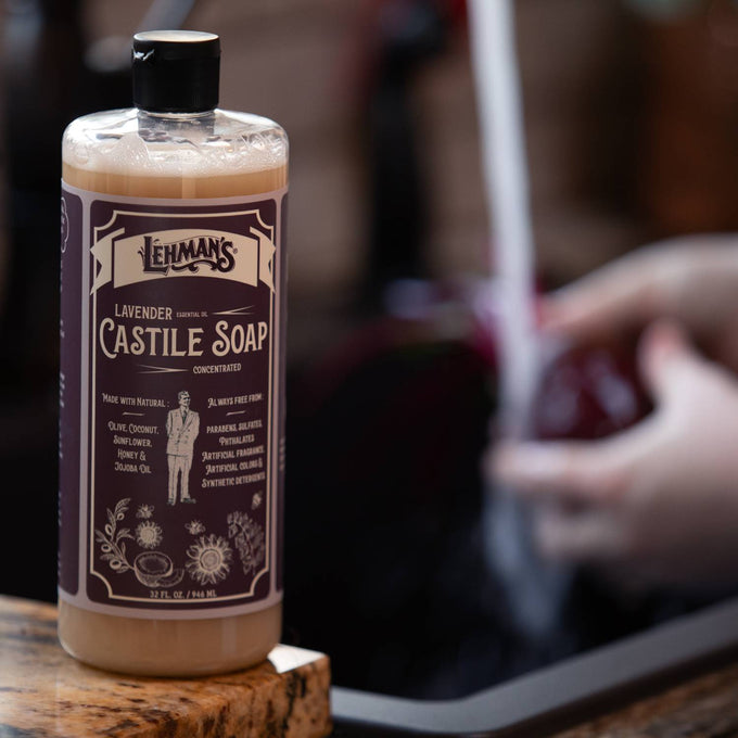 Amber bottle of Lehman's handcrafted lavender castile soap with natural plant oils, shown near a kitchen sink where produce is being rinsed, highlighting USA made soap for Health & Wellness and Grooming collections.