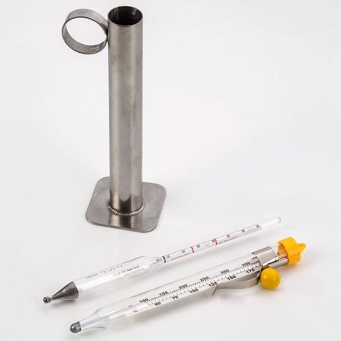 Glass encased thermometer and hydrometer with temperature and sugar scales, accompanied by a tin test cup holder, designed for maple syrup testing and farm tool collections.