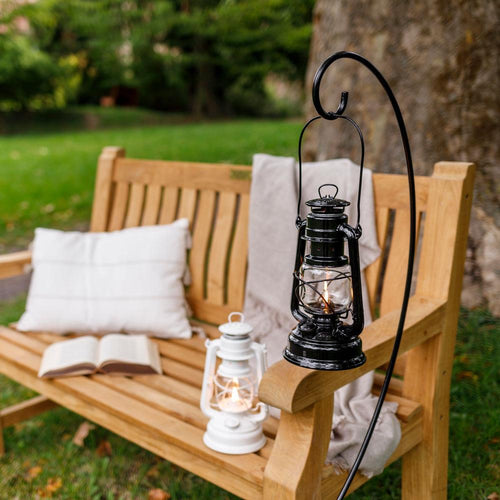 Black metal lantern holder with curved hook suspending a lit lantern outdoors on a wooden bench armrest, featuring a two-piece design for easy assembly, ideal for camping and emergency supplies.